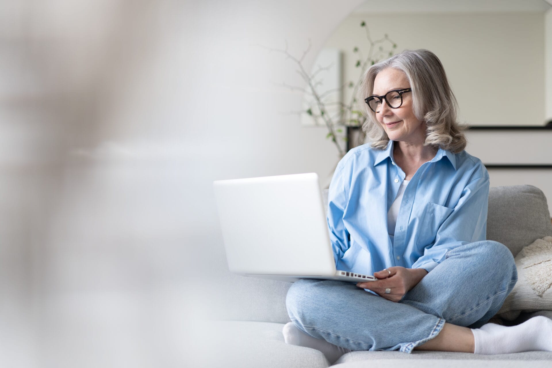 Smiling 50s mature woman sitting at home on sofa, holding laptop. Senior happy business woman using pc technologies, working, chatting, spending time in social media internet, ecommerce.